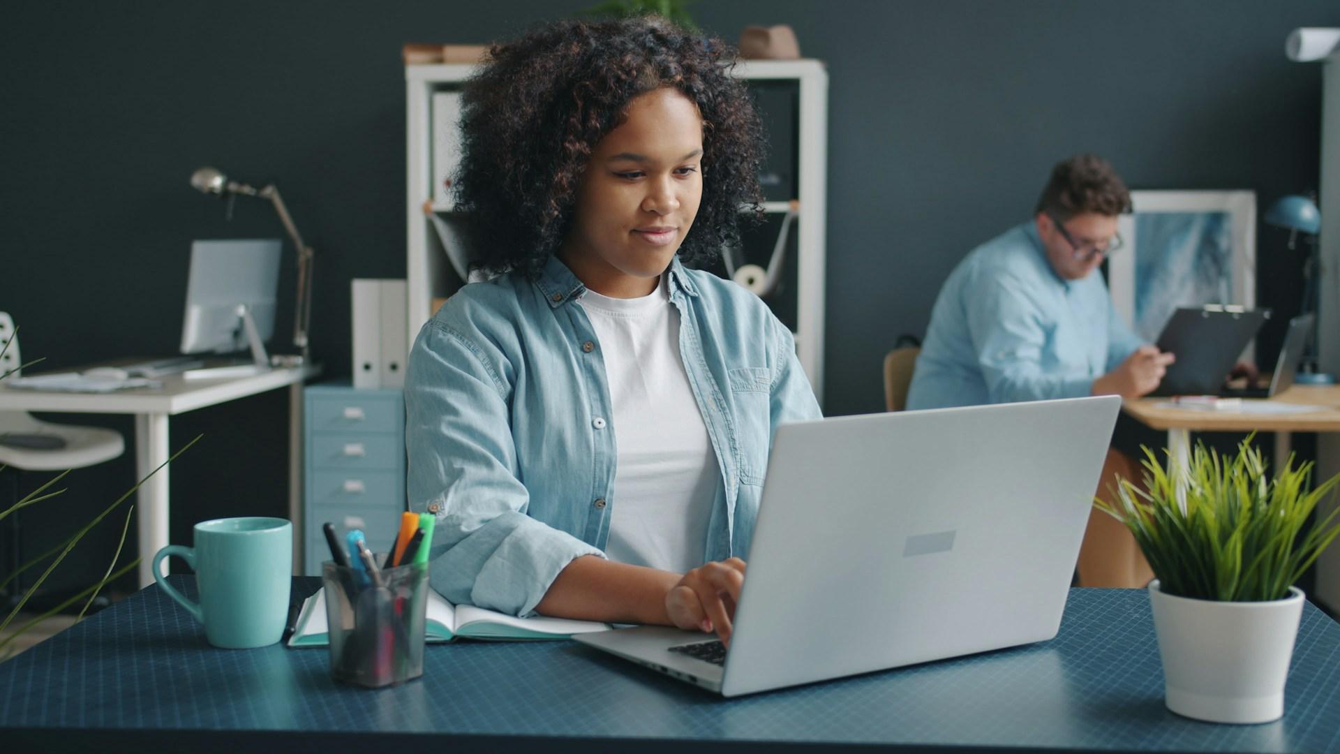 A person working at a laptop in a blue grey office, with a coworker in the background.