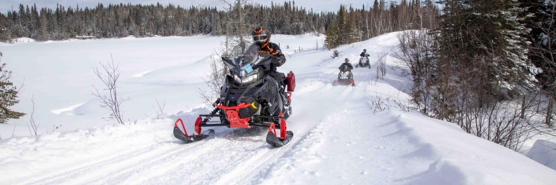 Snowmobiles riding on a snowy trail through a forest.